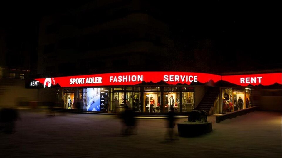 A shop with a bright red sign displaying "Sport Adler" and "Fashion Service". The scene is at night, and blurred figures can be seen in the foreground.