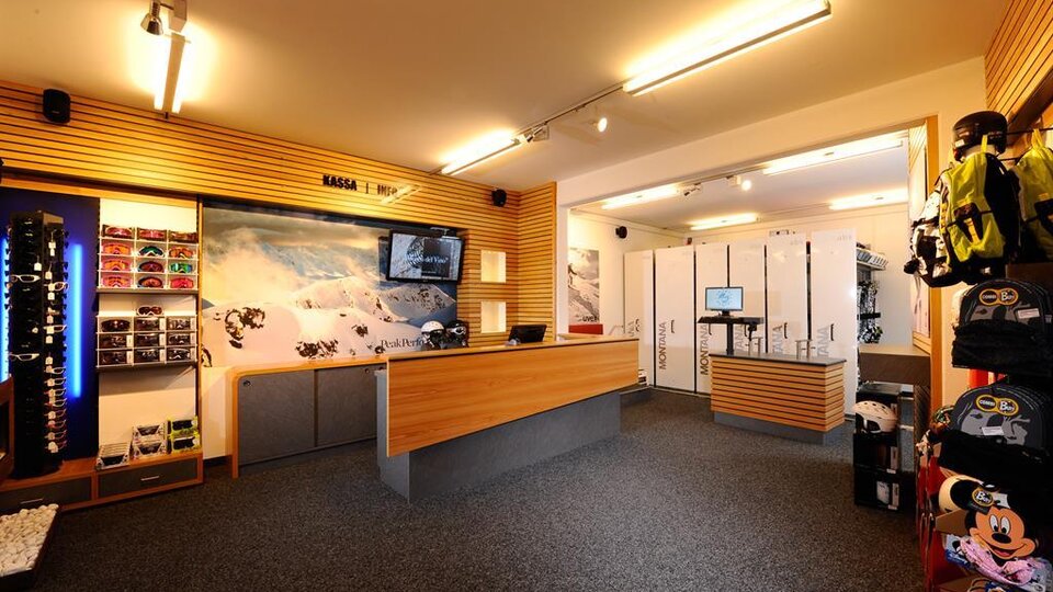 A modern rental space with wooden shelves and a reception desk. In the background, ski equipment and storage areas are visible.