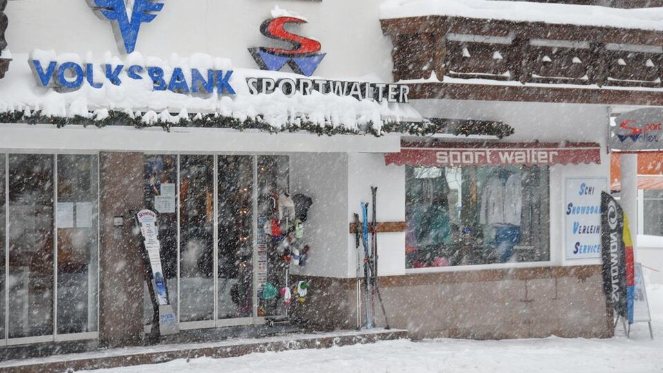 A sports store with ski equipment and a bank in winter. Thick snowflakes are falling and the surroundings are covered in snow.
