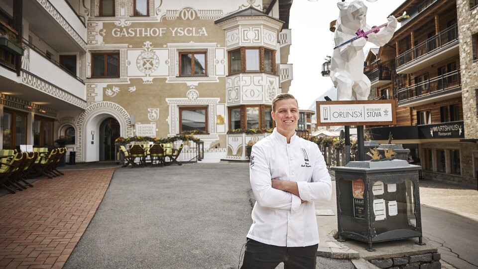 A chef stands smiling in front of the Yscla Inn. In the background, the impressive building with traditional details can be seen.