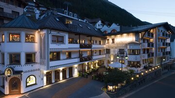 A beautiful hotel building in alpine style, surrounded by mountains. The facade is designed in soft colors and gently illuminated in the evening.