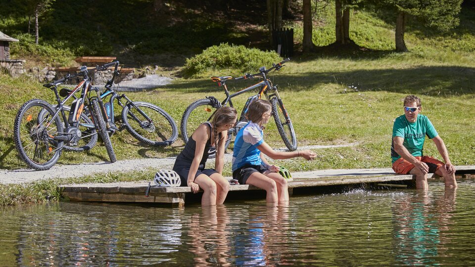 Two girls are sitting by the shore of a calm pond, dipping their feet in the water. In the background, there are bicycles standing among the trees. | © TVB Paznaun - Ischgl