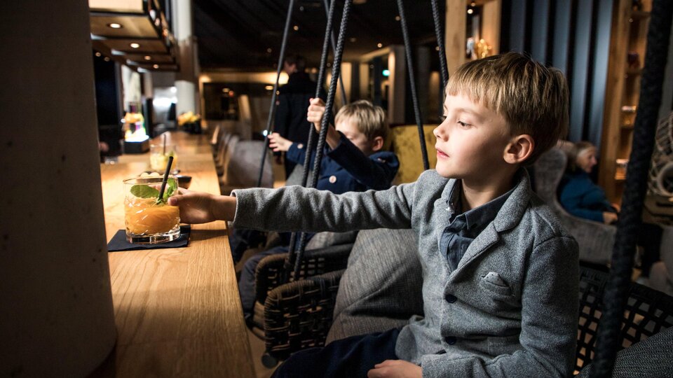 A boy in a plain gray suit is sitting at the bar and reaching for a drink. In the background, other children and a cozy interior are visible.