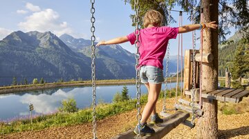 A child on a swing in a picturesque mountain landscape. In the background, a clear lake and tall mountains can be seen.