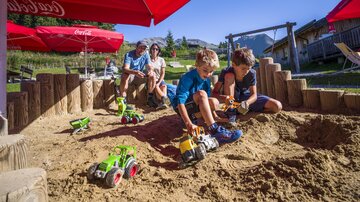Two children are playing with toy tractors in the sandbox. In the background, adults are sitting under red sun umbrellas.