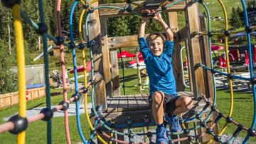 A cheerful boy is playing on a climbing frame made of wood and ropes. In the background, green meadows and play areas can be seen.