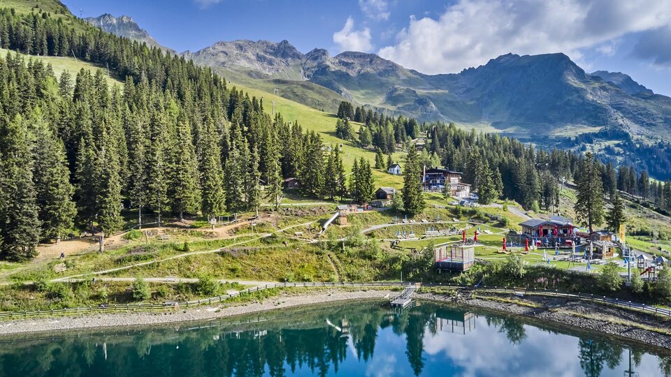 A picturesque mountain landscape with green forests and gentle hills. A calm lake reflects the surroundings.