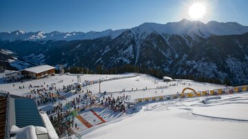 A wintry mountain landscape with skiers and a sunny sky. In the foreground, snow-covered slopes and a busy ski school can be seen.
