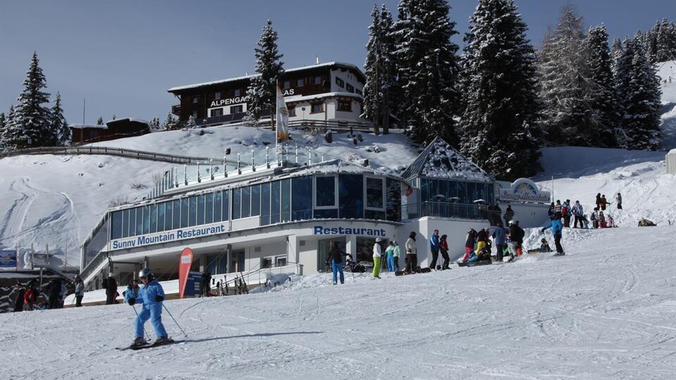 A snow-safe ski course with a modern restaurant in the foreground. Many people are actively enjoying winter fun in a picturesque mountain landscape.