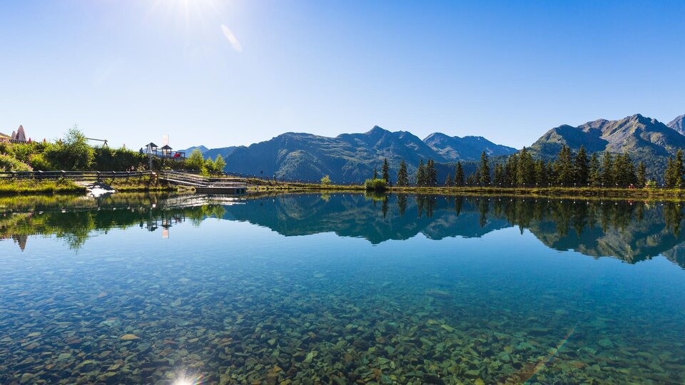A tranquil lake reflects the mountains and the blue sky. The surroundings are green and sunny.