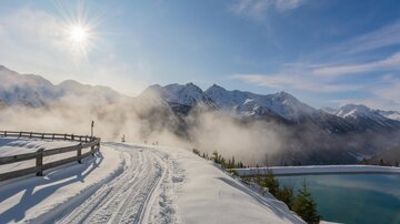 A snow-covered landscape with mountains in the background and bright sunshine. A path runs along a frozen pond.