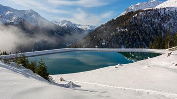 A clear mountain lake surrounded by snow-covered mountains. The sky is blue with a few clouds.