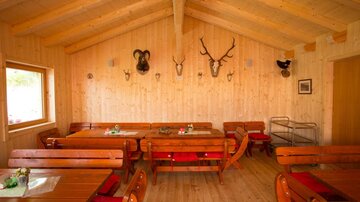 Ein gemütlicher Holzraum mit rustikalen Tischen und Bänken. An den Wänden hängen Jagdtrophäen und es gibt große Fenster, die für Tageslicht sorgen.
