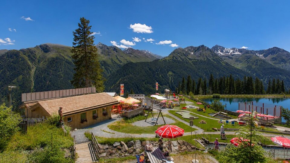 Eine alpine Freizeitanlage vor beeindruckender Bergkulisse: Im Vordergrund laden Holzterrasse, Liegewiese, Spielplatz und rote Sonnenschirme zum Verweilen ein, während rechts ein klarer Bergsee die umliegenden Gipfel spiegelt. Das helle Holzgebäude, die grünen Wiesen und der weite blaue Himmel vermitteln eine sommerliche, familienfreundliche Atmosphäre mitten in den Alpen.