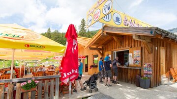 Ein gemütlicher Imbissstand aus Holz in der Natur. Es stehen Tische mit Sonnenschirmen und Besucher bestellen Essen.