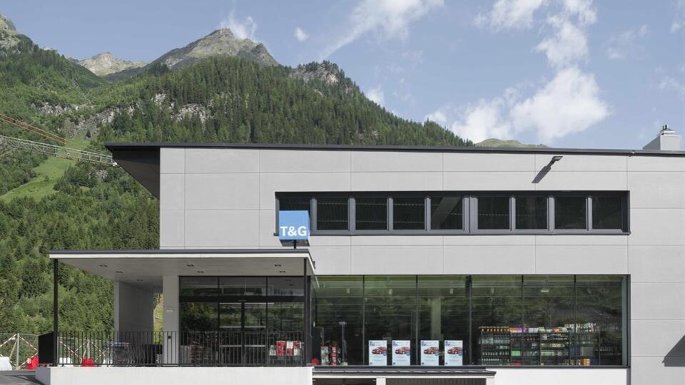 A modern building with large windows and a parking lot in front. In the background, green mountains are visible under a blue sky.
