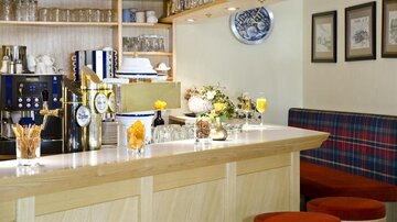 A cozy bar with wooden shelves and a coffee machine. Drinks and decorative elements are on the counter, while the seating is upholstered in red.