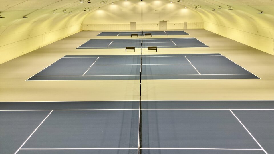 Eine moderne Tennishalle mit mehreren Spielfeldern. Die blauen Plätze sind gut beleuchtet und bieten Platz für mehrere Spiele.
