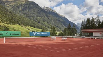 Ein Tennisspielplatz mit rotem Sand, umgeben von malerischen Bergen und grünen Bäumen. Der Himmel ist bewölkt, aber die Landschaft ist beeindruckend.