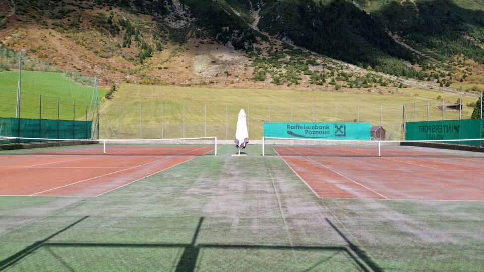 A tennis court in a picturesque mountain landscape. The surroundings are green and sunny with a clear blue sky.