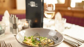 A stylishly set table with a plate of pasta and fresh vegetables. In the background, there is a drink and a menu card visible.