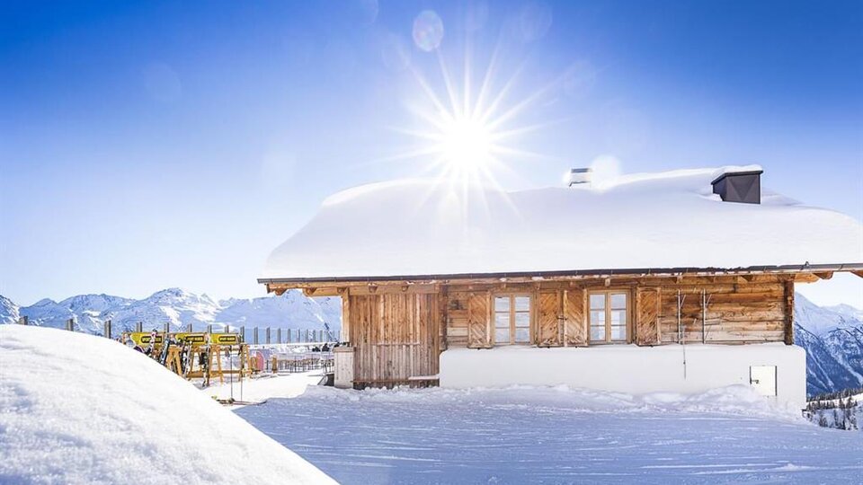 A charming wooden house in the snow with bright sunlight. Snow-covered mountains are visible in the background.