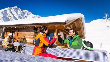 A couple enjoys drinks in a snowy area in front of a mountain hut. In the background, there are snow-covered mountains and more visitors.