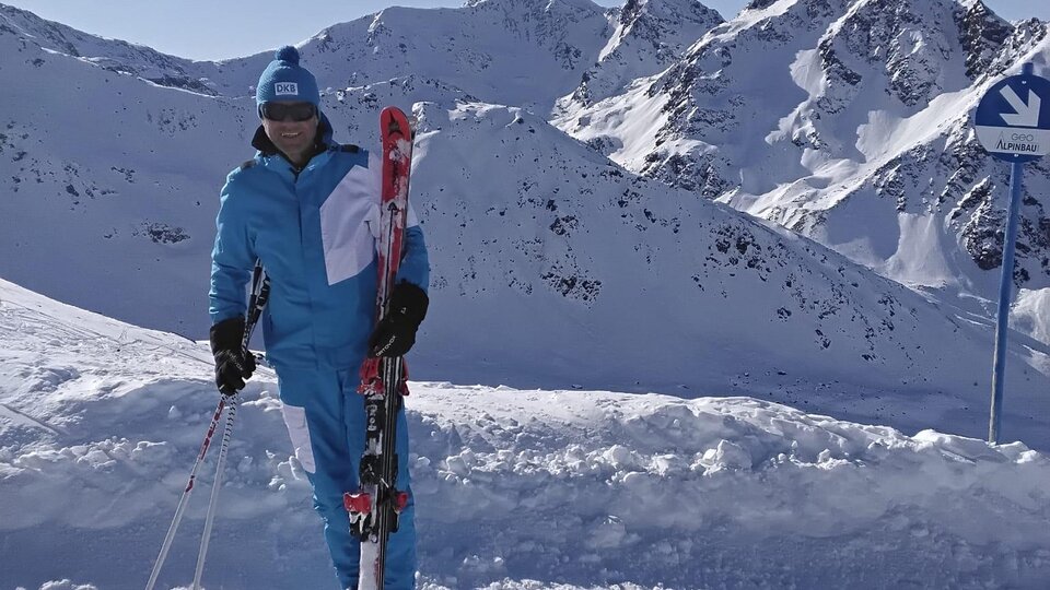 Eine Person steht im Schnee mit Skiern in der Hand. Im Hintergrund sind schneebedeckte Berge und ein klarer blauer Himmel zu sehen.