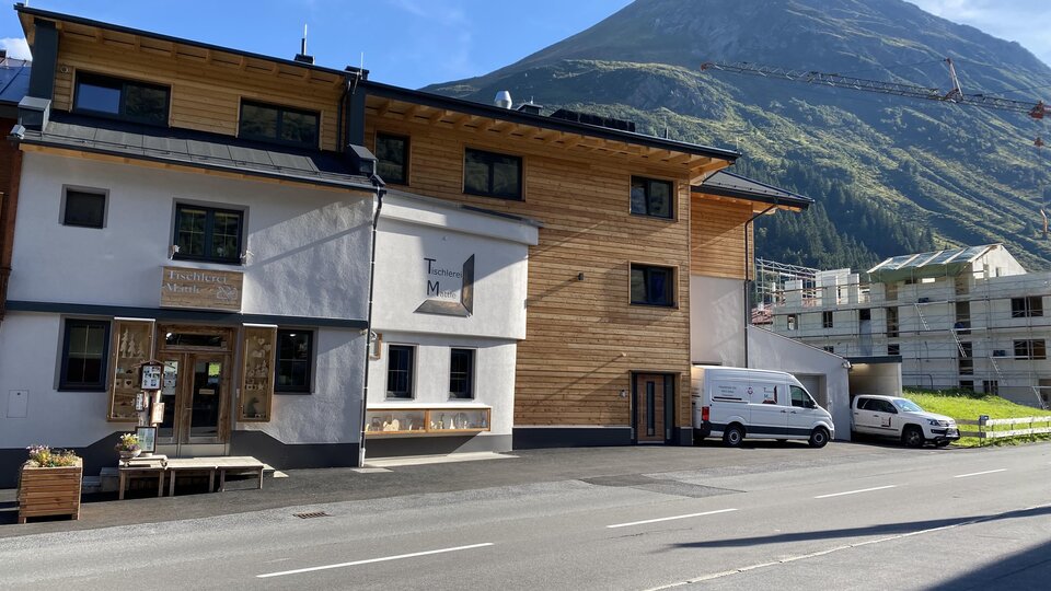 A modern building with wood cladding and large windows in a mountain landscape. In the background, a tall mountain is visible under a blue sky.