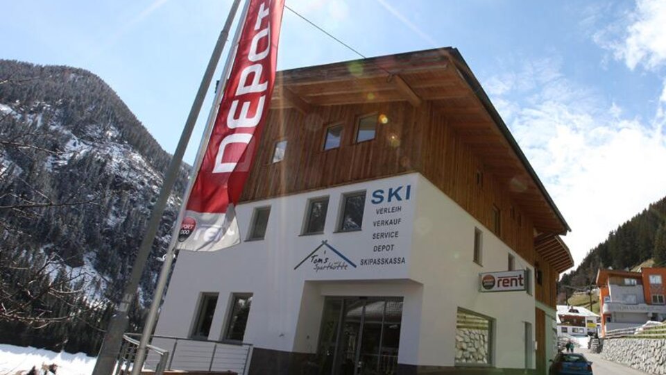 A modern shop with a wooden facade that offers ski rental and sales. The surroundings are mountainous and snow-covered.