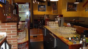 A cozy bar with a long counter and many glasses. In the background, there are tables and chairs for guests.