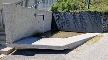 A modern fountain made of concrete with a water flow and shallow basin. In the background, green shrubs can be seen.