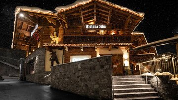 A traditional, well-lit mountain house in a wintery setting. The entrance is inviting and atmospheric snowflakes fall gently.