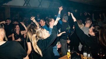A lively party scene with dancing people in a dark room. The atmosphere is energetic, and many are holding drinks.