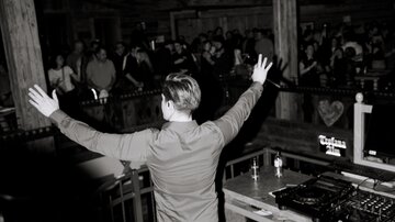 A DJ stands behind the booth and raises his arms while the audience cheers him on. The scene is darker, with atmospheric lighting and a lively atmosphere.