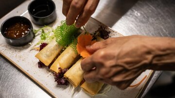A plate with spring rolls is being decorated. There are also various sauces and fresh vegetables.