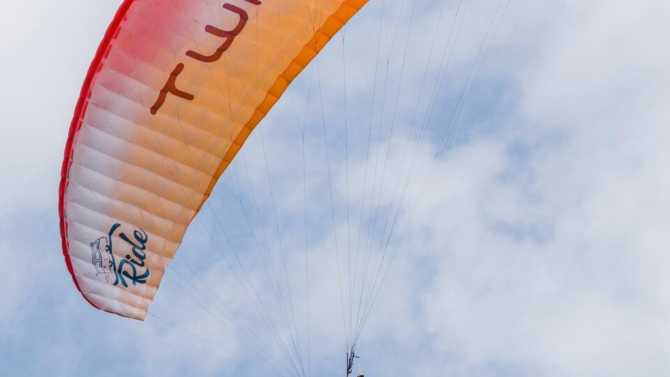 A paraglider hovers high in the sky. The canopy is orange and red, and the clouds are barely visible.