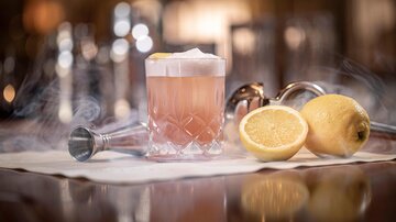 An elegant cocktail in a glass chalice with a slice of lemon. Next to it, there are two halved lemons on a white cloth.