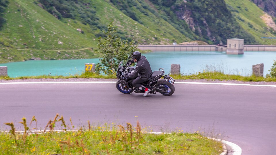 A motorcyclist rides along a winding road next to a clear lake. The green landscape around him creates an impressive backdrop.