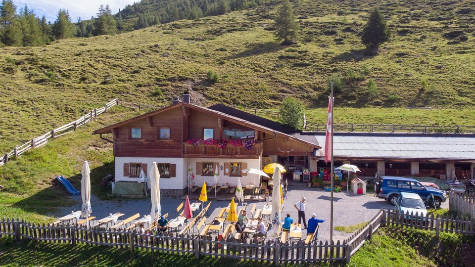 An Alpine inn with a terrace and deck chairs, surrounded by green meadows. In the background, gentle hills and trees rise up.