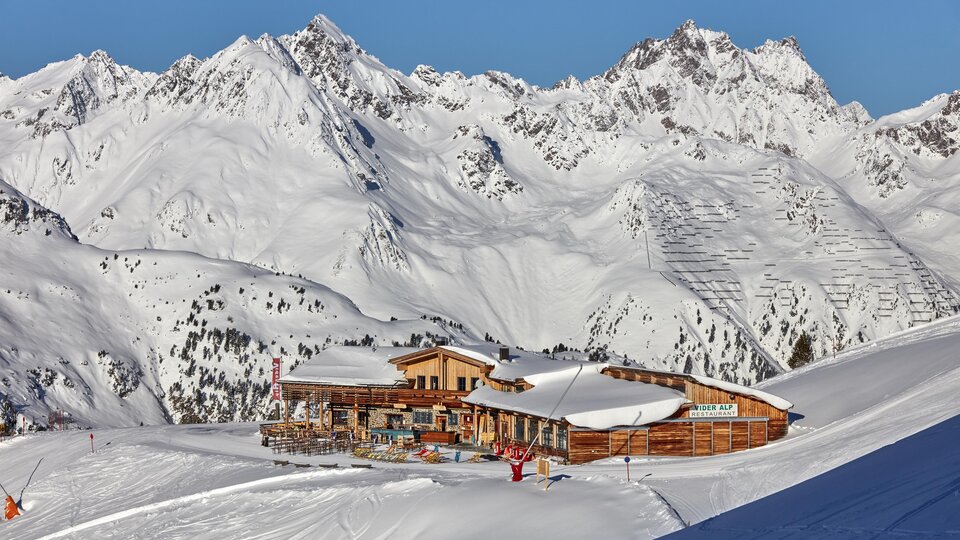 A cozy mountain cabin is situated in a snowy landscape. In the background, majestic mountains rise up under a clear sky.
