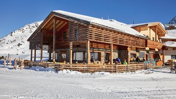 A cozy wooden building in the snow, surrounded by mountains. There are people sitting outside enjoying the winter landscape.