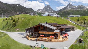 A cozy mountain cabin surrounded by green hills and impressive mountains. The cabin has a terrace with red and white sun umbrellas.