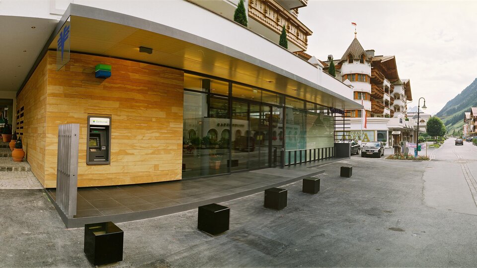 A modern building with large windows and wooden cladding. In the background, traditional architecture and mountains are visible.