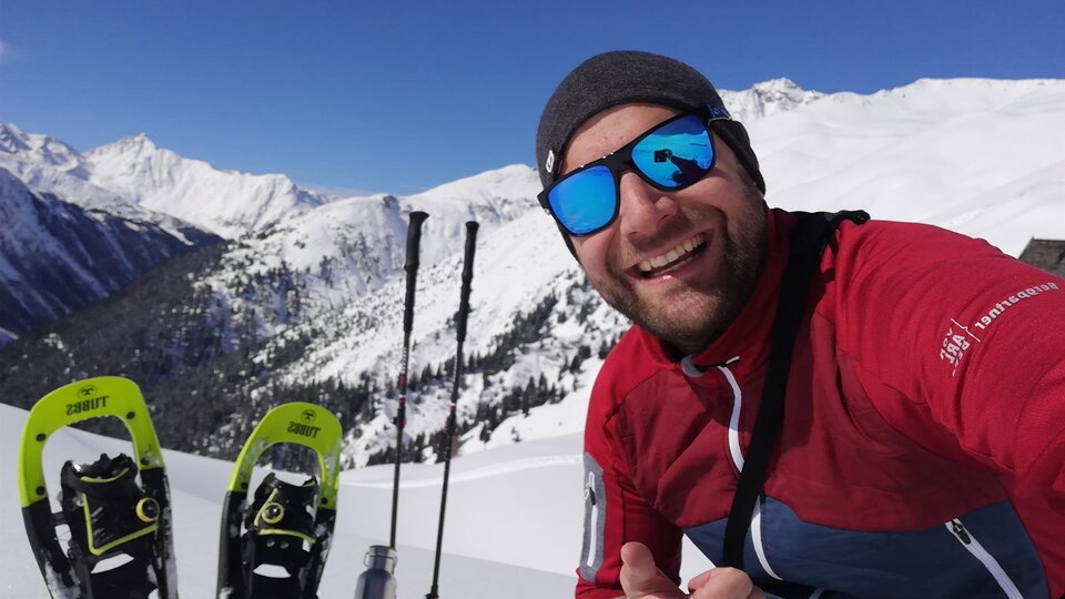 A man takes a selfie in the snowy mountains. In the background, snowy peaks and snowshoes can be seen.