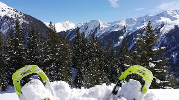 Snowshoes stand in fresh snow, surrounded by a picturesque mountain landscape. In the background, high mountains and a clear blue sky are visible.