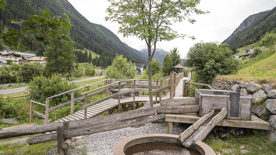A quiet landscape with a wooden walkway and a water hole. Surrounded by green hills and trees.