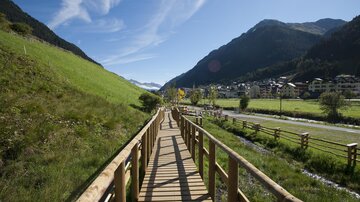 A beautiful wooden walkway leads through a green landscape with mountains in the background. The weather is clear and nature radiates tranquility.