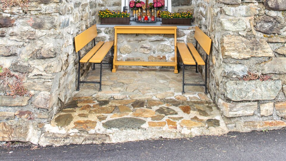 A small, rustic chapel made of stone and wood stands in a green environment. In front of the chapel, two benches and flowers can be seen. | © Bernhard Gruber – Photography