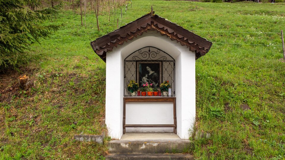 A small chapel on a green hill. In front of the chapel stands a window with flowers and a picture.